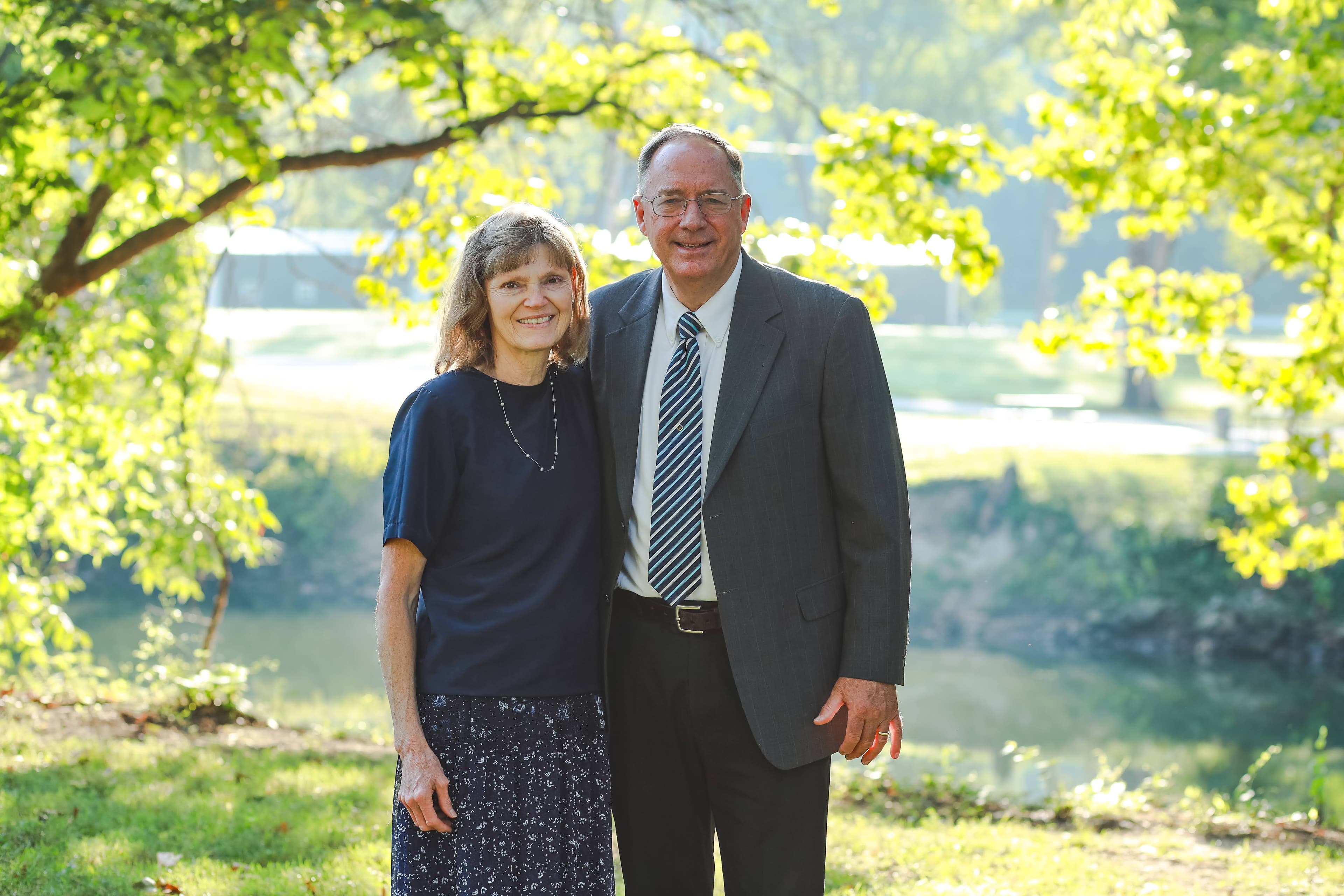 Smiling older couple posing together in a sunny park with lush green trees and water.