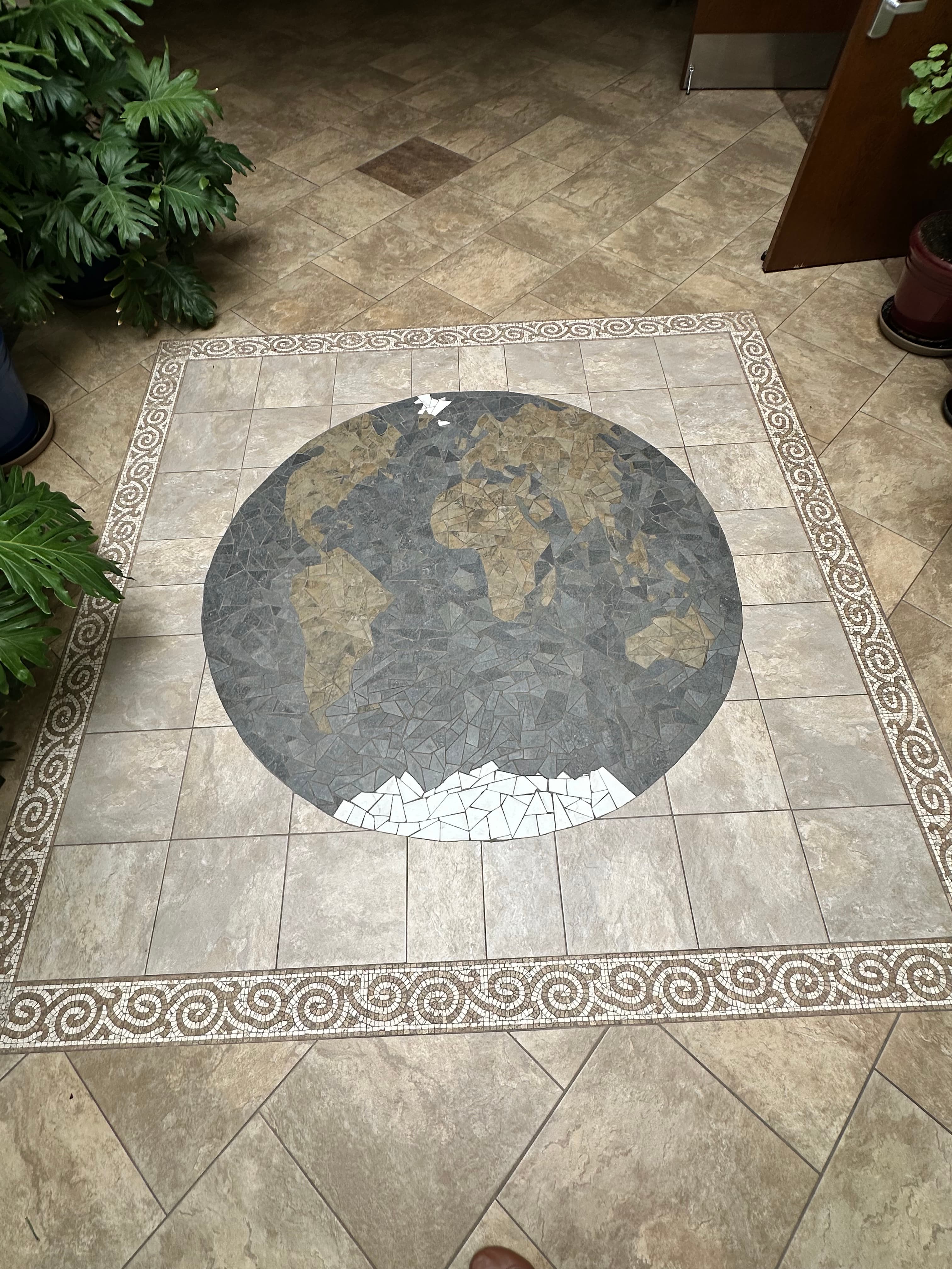 Circular mosaic world map on tiled floor with tan continents, gray oceans, and white Antarctica.
