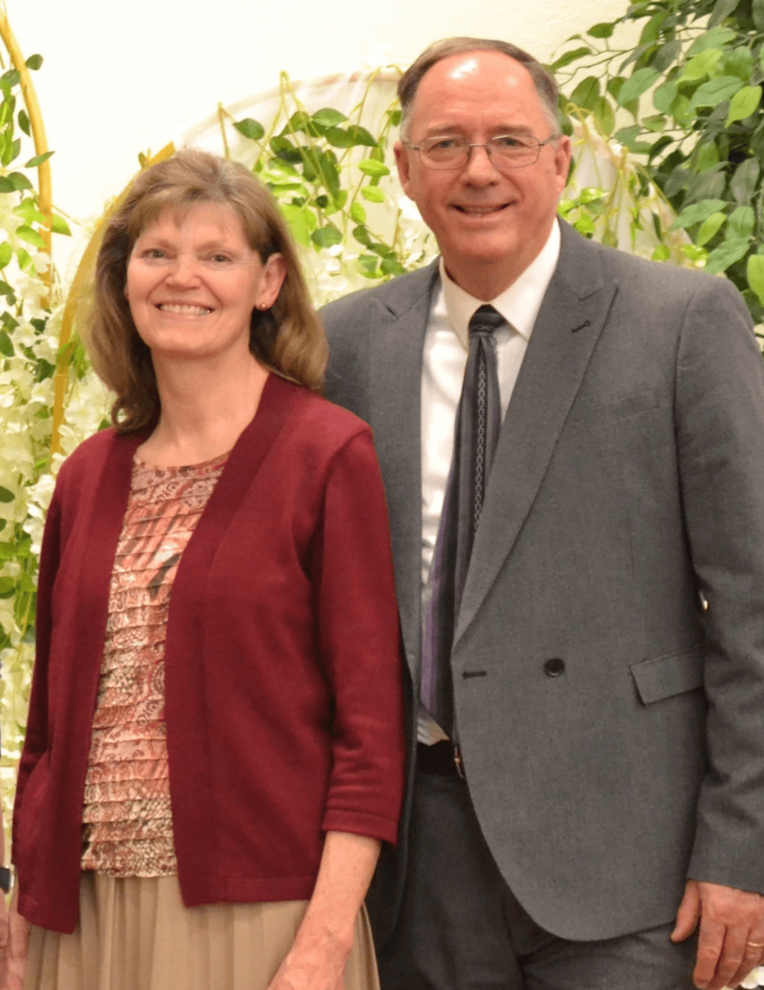 Smiling woman in maroon cardigan and man in grey suit standing before a floral backdrop.