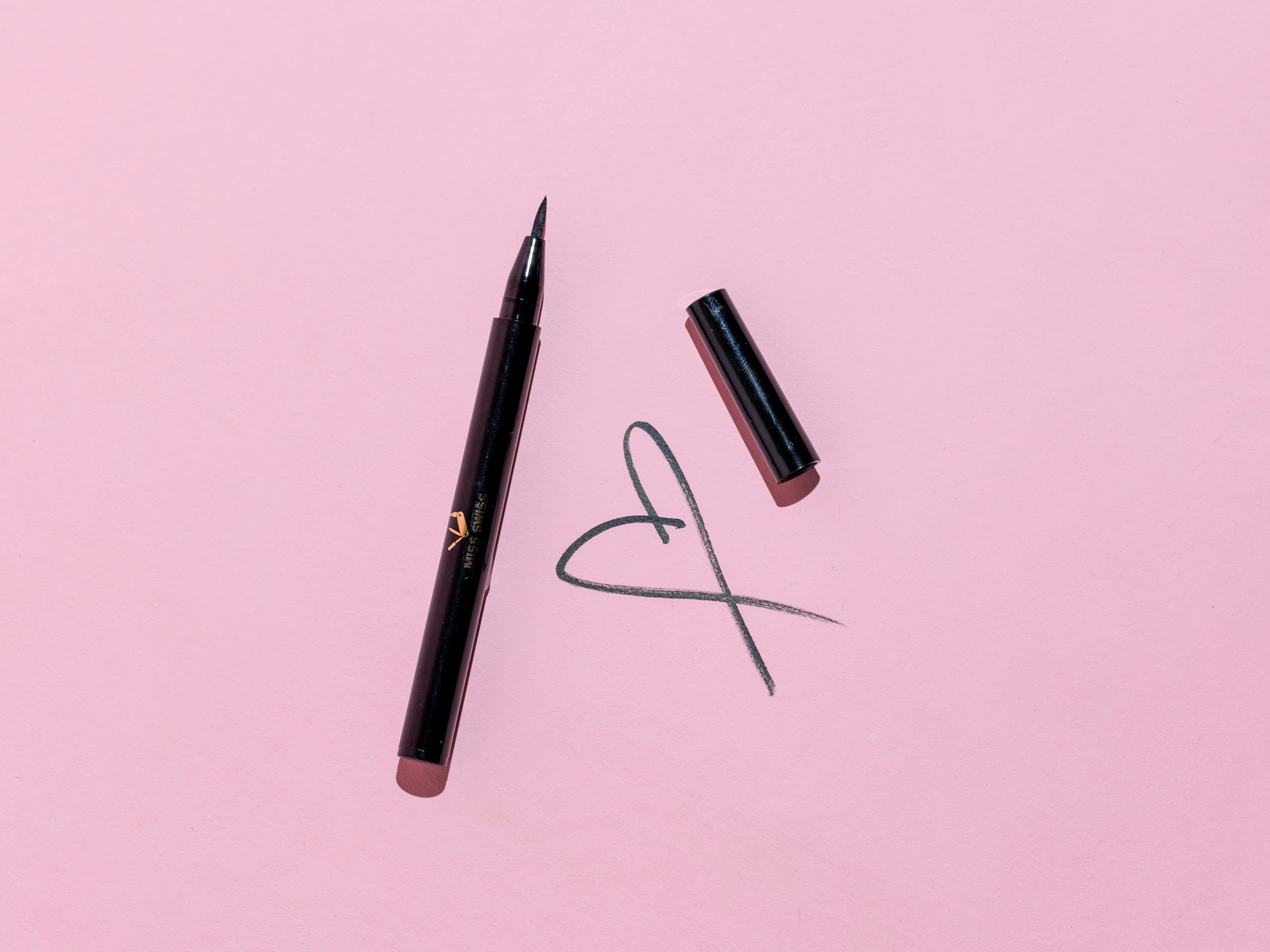 Black liquid eyeliner pen and cap on a pink background with a hand-drawn heart.